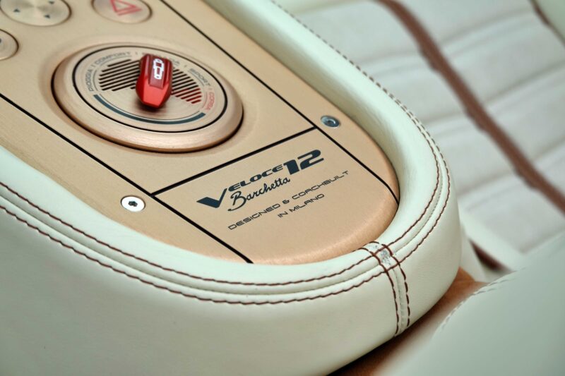 Close-up of a car’s interior control panel featuring a red switch, round dials, and a “Veloce 12 Barchetta” plaque with tan and cream leather upholstery in the Touring Superleggera Debuts New Veloce12 “Barchetta” Restomod.