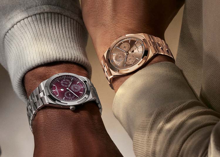 Two wrists display luxury timepieces: a Vacheron Constantin silver watch with a burgundy face on the left, and an Overseas Perpetual Calendar gold-tone watch with a light-colored face on the right.