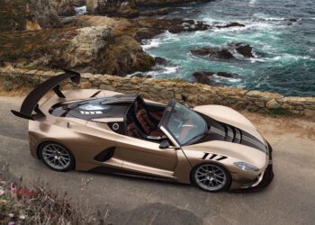 A gold Hennessey Venom F5 convertible with black accents is parked on a coastal road overlooking rocky cliffs and the ocean.