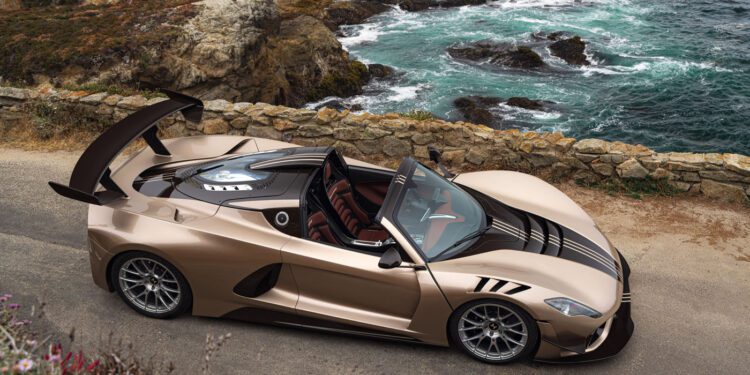 A gold Hennessey Venom F5 convertible with black accents is parked on a coastal road overlooking rocky cliffs and the ocean.
