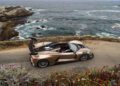 A gold Hennessey Venom F5 sports car with a large rear wing—boasting 2,031HP—is parked on a coastal road overlooking rocky cliffs and the ocean.