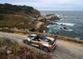 A gold Hennessey Venom F5 with a manual transmission and a large rear wing is parked on a coastal road next to rocky cliffs and the ocean under a cloudy sky.