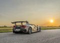 A silver Hennessey Venom F5 sports car with a large rear wing is parked on a paved road at sunset, surrounded by grassy fields and trees in the background.