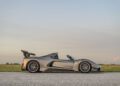 A silver Hennessey Venom F5 convertible sports car with a large rear spoiler is parked on a paved road next to a grassy field under a clear sky.