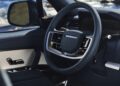 A close-up of a Range Rover SV’s steering wheel and dashboard, showing modern controls, with an ocean view near Asilomar visible through the window—perfect for Monterey Car Week enthusiasts.