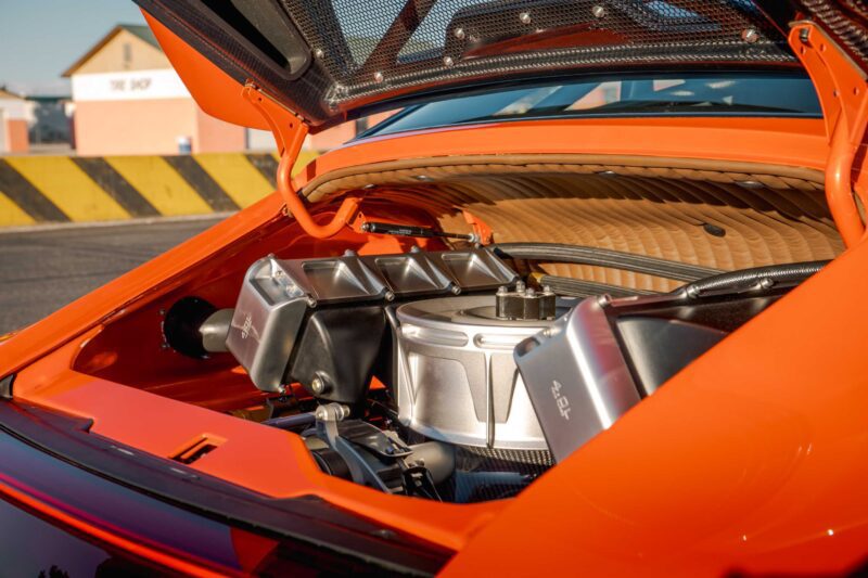 A close-up view of the engine bay of an orange Gunther Werks Turbo Tested sports car with the rear hood open, showcasing the engine and mechanical components—bar none, the gnarliest Porsche restomod yet.