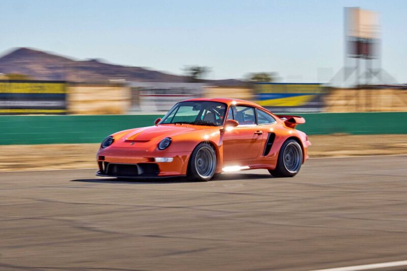 An orange Gunther Werks Turbo Tested sports car speeds along a racetrack with blurred background, indicating motion; mountains and advertising banners are visible in the distance, showcasing the gnarliest Porsche restomod yet.