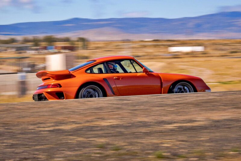 A Gunther Werks Turbo Tested: Bar None The Gnarliest Porsche Restomod Yet, this orange sports car with a rear spoiler speeds along a paved road through a dry, open landscape, framed by distant mountains and blue sky.