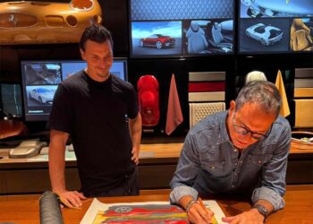 Two men are in a design studio; one sketches a car on paper while the other observes. Behind them, images of the Ferrari F80 and materials are displayed—echoing the creative spirit of Flavio Manzoni.