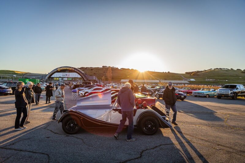 People gather around classic and sports cars at an outdoor car show during sunrise, with a Continental arch and hilly landscape in the background, celebrating Monterey Car Week 2025 presented by duPont REGISTRY.