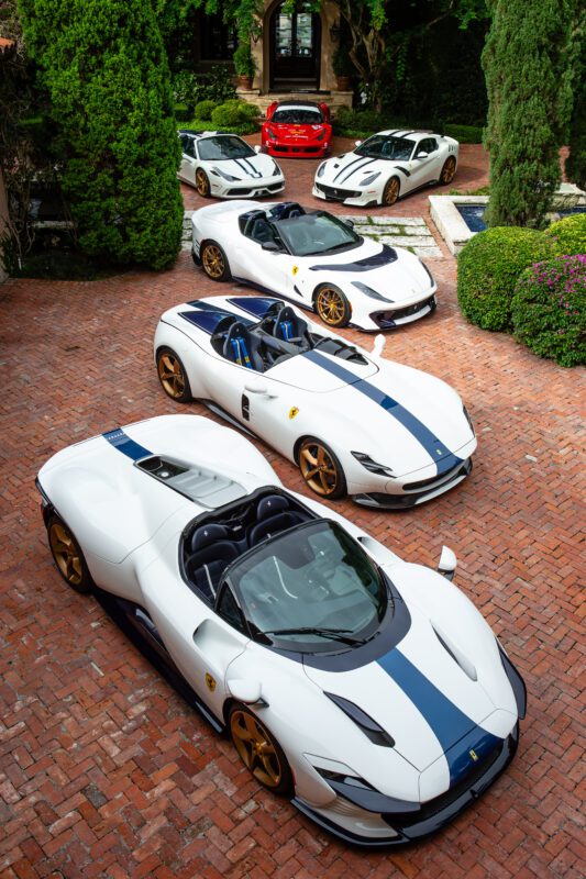 Five luxury sports cars, mostly white with blue racing stripes and gold wheels, are parked on a brick driveway near greenery—an impressive display worthy of the NART Collection or any fine art enthusiast’s dream.