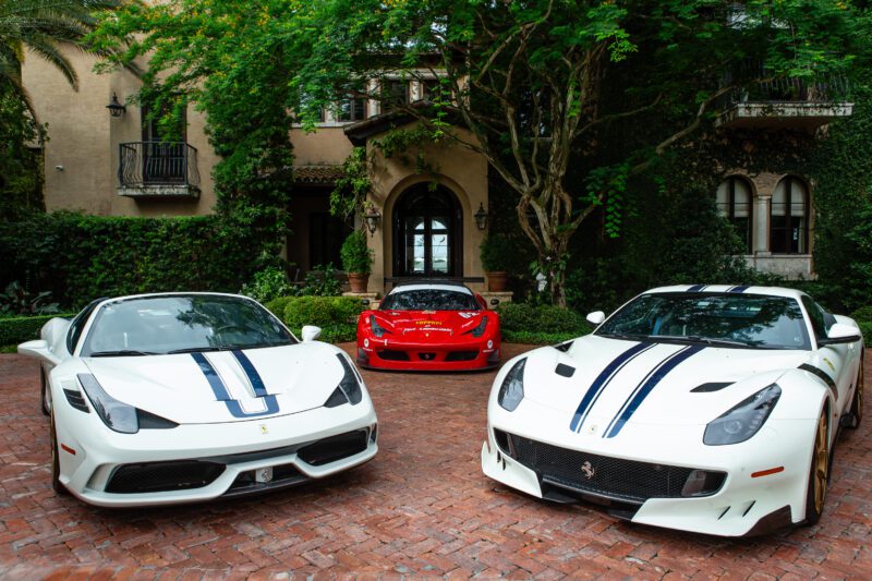 Three sports cars, part of the Fine NART Collection—two white with blue racing stripes and one red—are parked on a brick driveway in front of a large house surrounded by greenery.