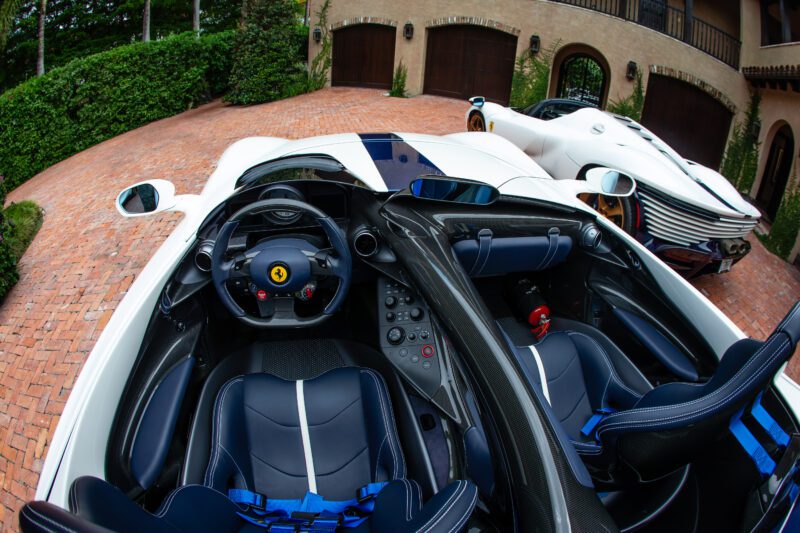 Fisheye view of a white Ferrari from the Fine NART Collection with black-and-blue interior, parked on a brick driveway, with another white sports car in the background in front of a garage.