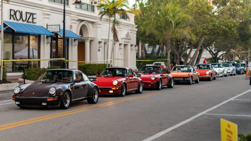 A row of vintage Porsche cars is parked along a street in front of shops and trees on a clear day, capturing the spirit of Naples Cars on 5th.