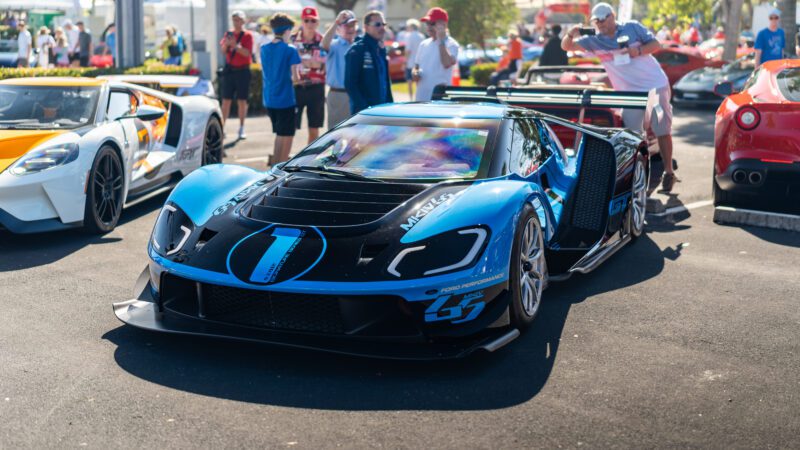 A blue and black race car with number 67 displayed on the hood is parked at the Naples Cars on 5th show, surrounded by people and other sports cars.