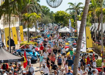 A large crowd walks among rows of parked sports cars at the 2026 Concours Naples Cars on 5th, an outdoor car show lined with palm trees, vendor tents, and banners on a sunny day.