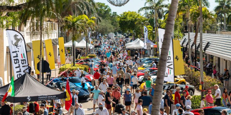 A large crowd walks among rows of parked sports cars at the 2026 Concours Naples Cars on 5th, an outdoor car show lined with palm trees, vendor tents, and banners on a sunny day.