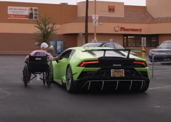 Garet Thomas, in a wheelchair, moves toward a green Lamborghini parked in a pharmacy lot during Monterey Car Week 2025. A "We're Hiring" sign is visible on the building in the background.