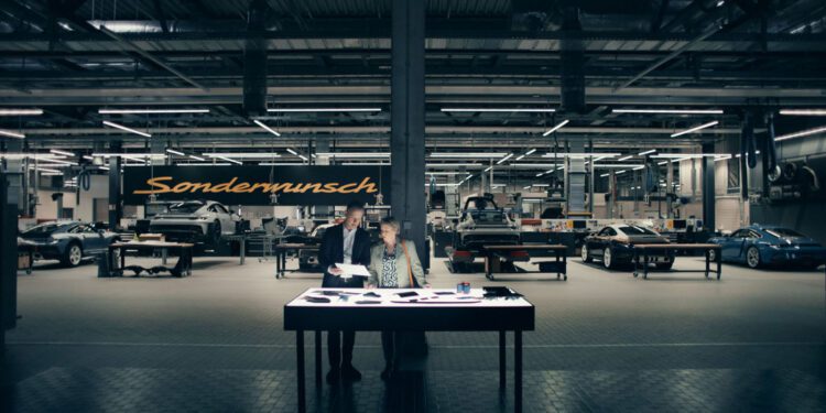 Two people stand at a table reviewing documents in a well-lit Porsche workshop, with several cars and a large "Sonderwunsch" sign in the background, capturing a behind-the-scenes moment from the video series.