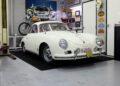 A white vintage Porsche 356 coupe is parked indoors on a black mat, surrounded by automotive memorabilia and classic car decor.
