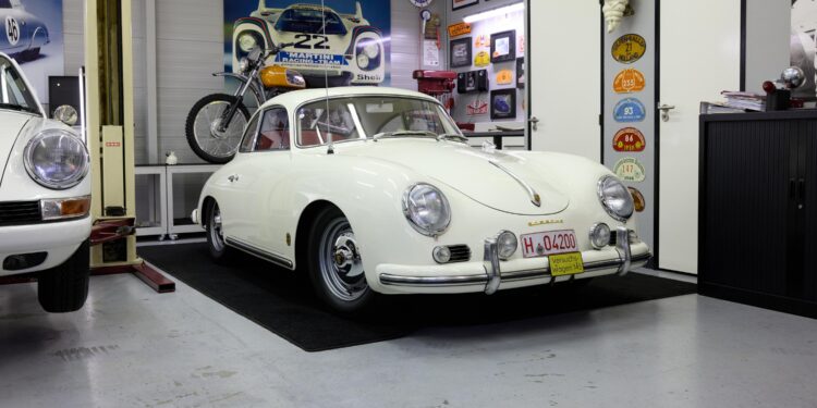 A white vintage Porsche 356 coupe is parked indoors on a black mat, surrounded by automotive memorabilia and classic car decor.