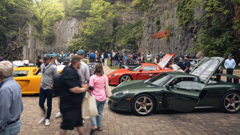 People gather around several sports cars, including a yellow Carrera GT, on a stone courtyard surrounded by rocky cliffs and greenery, enjoying a 2026 Porsche Plática event.