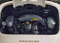 View of a classic Porsche engine compartment with visible air filters, wiring, and engine components, taken from behind with the rear hood open. The Porsche logo is on the lower edge.