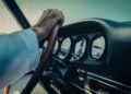 Close-up of a person's hand on a wooden steering wheel, with classic Porsche dashboard dials and controls in the background.
