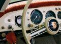 Close-up of a vintage Porsche dashboard featuring cream steering wheel, analog gauges, ignition key, and textured red-brown dashboard trim.