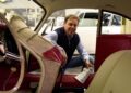 A man kneels outside a classic car, cleaning the interior with a towel. The car has red and beige upholstery, and another car is visible through the open door.