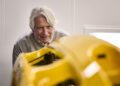 An older man with gray hair closely inspects a yellow Porsche part, possibly for a Sonderwunsch project, in a well-lit industrial setting.