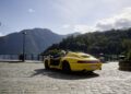 A yellow Porsche convertible, part of the Sonderwunsch video series, is parked on a cobblestone street near a lake, framed by tree-covered mountains and a cloudy sky in the background.