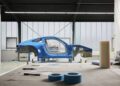 A blue Porsche chassis is positioned in the center of a workshop, surrounded by windows; rolls of tape and tools are visible on a table in the foreground, capturing the essence of a Sonderwunsch project from the video series.