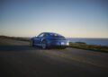 A blue Porsche sports car drives along a coastal road at sunset, with the ocean and horizon visible in the background—part of an exclusive Sonderwunsch video series.