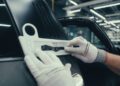 A person wearing white gloves applies a "Targa" badge to the rear side of a black Porsche in a well-lit workshop, capturing a Behind The Scenes moment of meticulous craftsmanship.