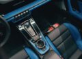 Overhead view of a modern Porsche interior with blue accents, showing the center console, gear shifter, cup holder, and a red fire extinguisher on the floor—perfect for a Sonderwunsch video series.
