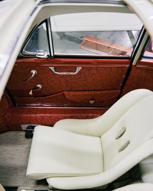 A close-up view of a vintage car’s interior, showing a cream-colored seat, red patterned door panel, and beige carpeted floor.