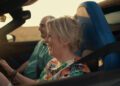 A woman drives a Porsche convertible with a man in the passenger seat, both smiling and wearing seatbelts, enjoying daylight—perfect for a Sonderwunsch video series.