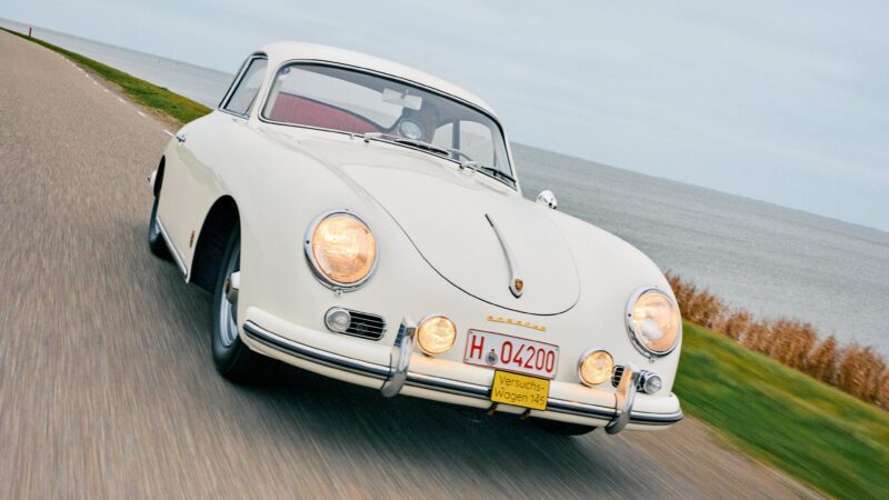 A classic white Porsche 356 coupe driving on a coastal road with headlights on and a German test license plate visible.
