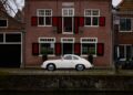 A vintage white Porsche 356, possibly a Secret One-Off by Henk Spin, is parked on a cobblestone street in front of a brick building with red and black shutters.