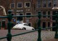 A white vintage Porsche 356 is parked on a cobblestone street in front of old brick buildings, partially obscured by a green metal fence—a secret one-off envisioned by Henk Spin.