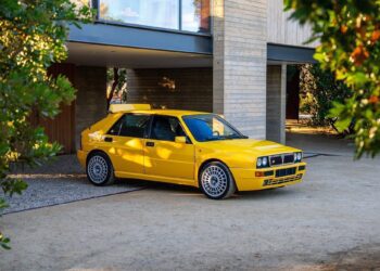 A yellow Lancia Delta Integrale hatchback is parked on a driveway in front of a modern house, surrounded by trees and sunlight—a true Petrolicious moment.
