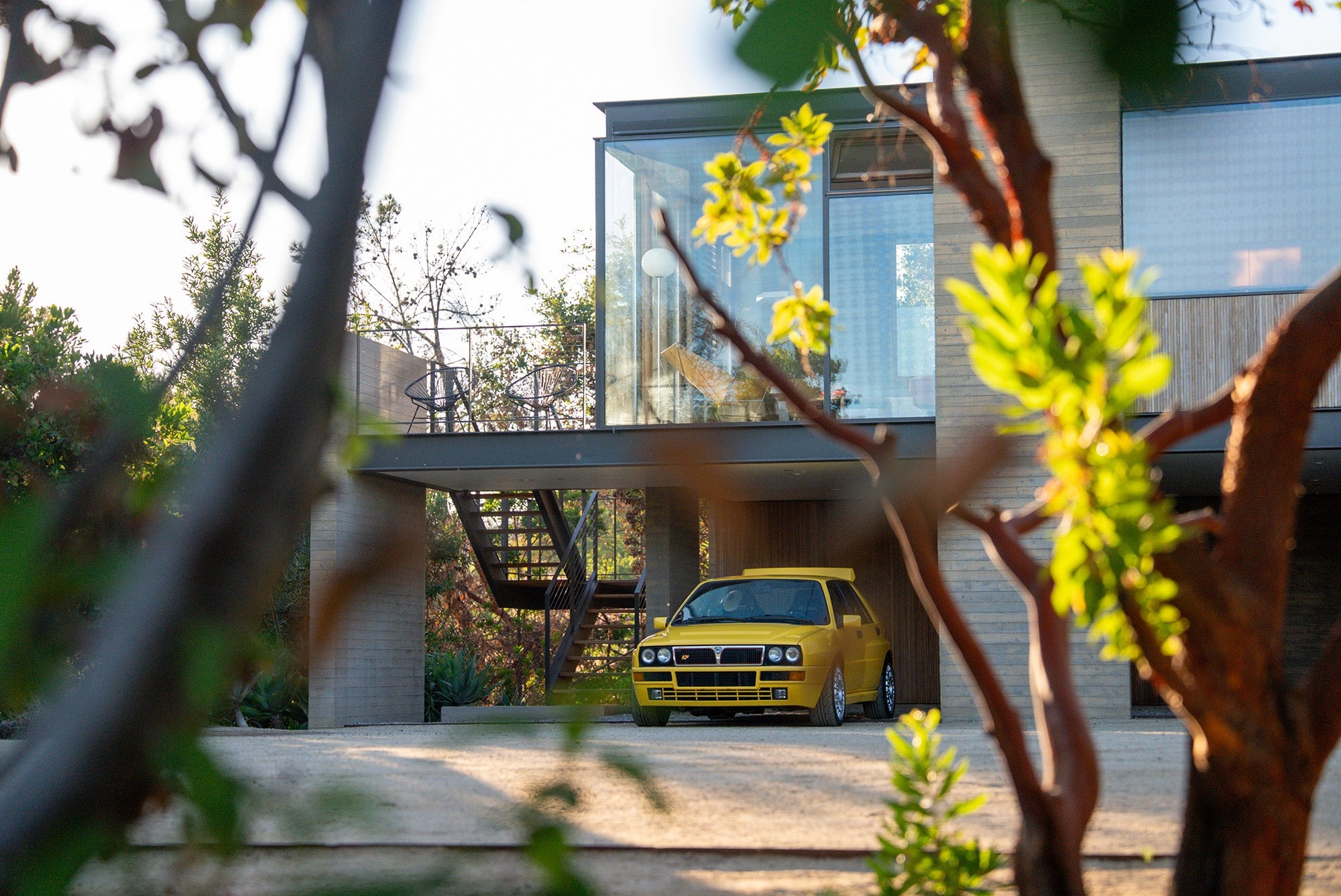 A yellow Lancia Delta Integrale Evo 2 is parked in the driveway of a modern two-story house, its bold lines partially obscured by tree branches in the foreground—a scene worthy of Petrolicious.