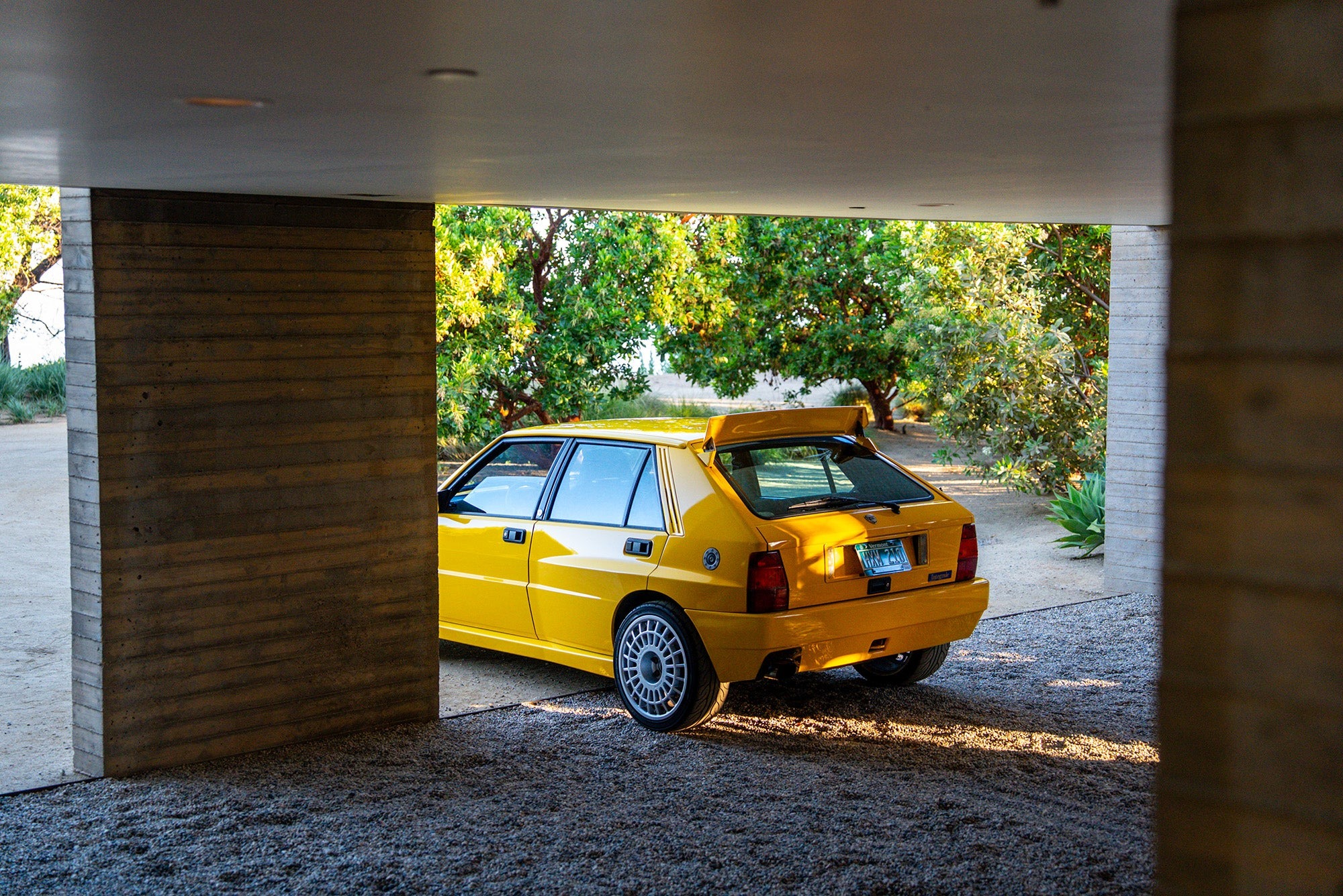 A yellow Lancia Delta Integrale hatchback with a rear spoiler is parked partially under a modern concrete carport, with trees and sunlight in the background—a true Petrolicious moment.
