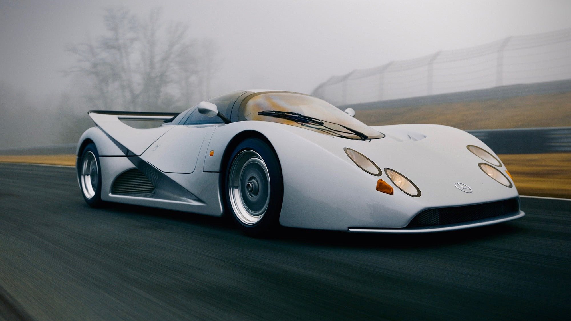 A white Lotec C1000 sports car with a low, aerodynamic design drives on a racetrack in foggy conditions, blurred trees and fencing in the background—perfect for a Petrolicious Film Friday feature.