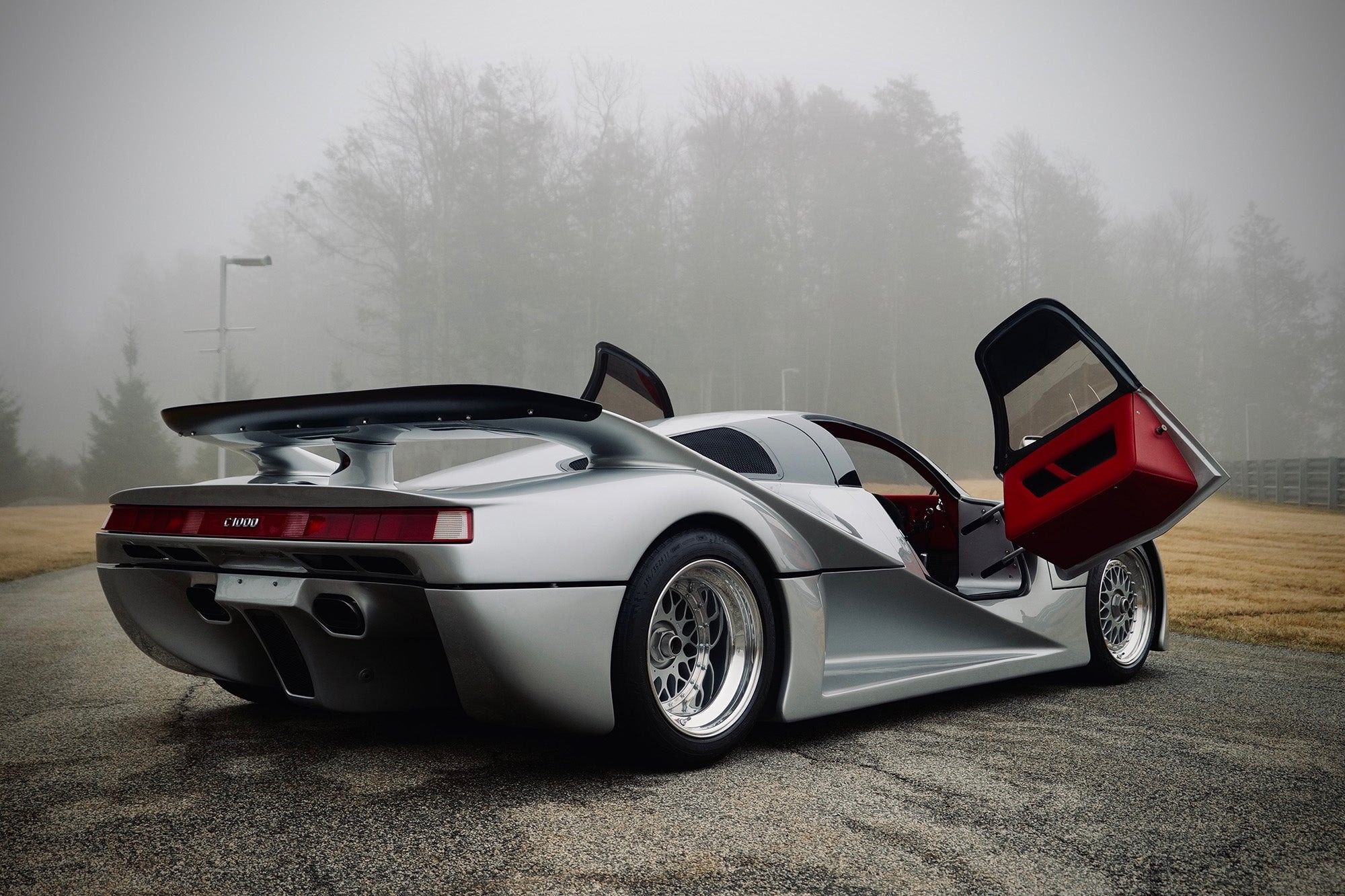 A silver Lotec C1000 sports car with its driver-side door open is parked on a foggy road, surrounded by trees and grass—a scene worthy of Petrolicious or Film Friday.