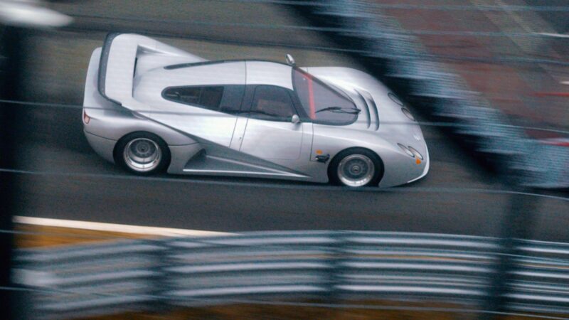 A silver Lotec C1000 sports car is driving at high speed on a racetrack, with blurred barriers and surroundings indicating motion—a true Film Friday moment for any Petrolicious enthusiast.