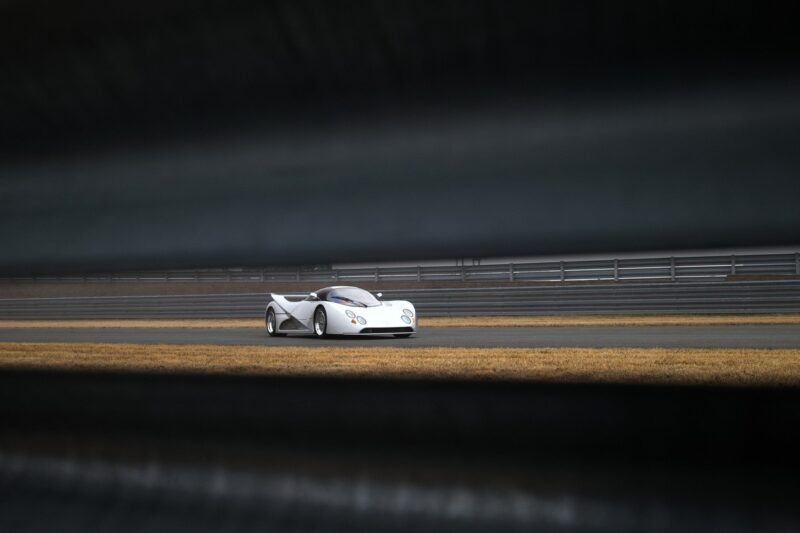 A white Lotec C1000 sports car is driving on a racetrack, viewed through metal railings with dry grass in the foreground—a scene worthy of Petrolicious Film Friday.