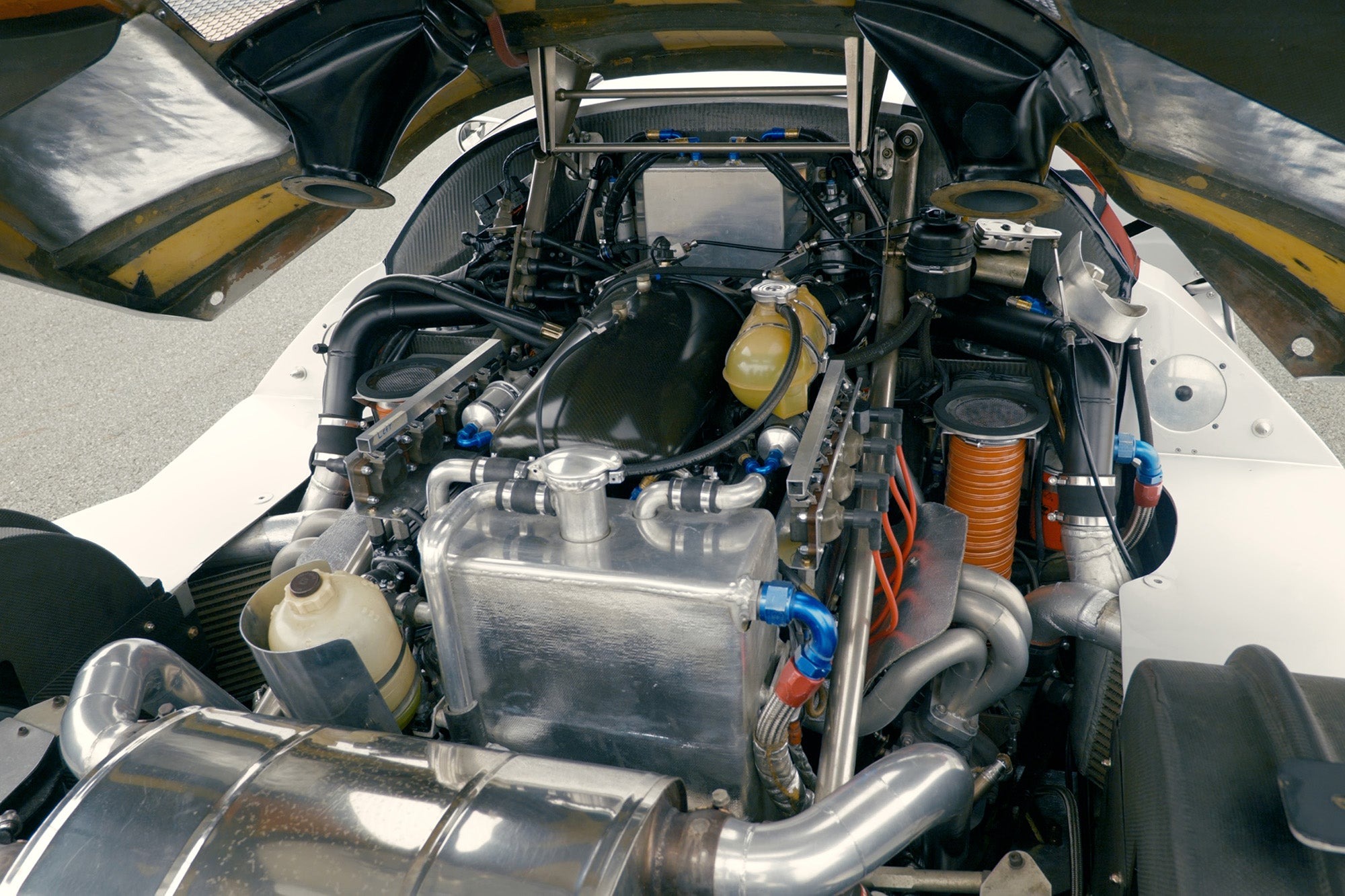 Close-up view of a Lotec C1000 race car’s engine bay featuring various mechanical components, pipes, and wires under an open rear hood—perfect for a Petrolicious Film Friday spotlight.