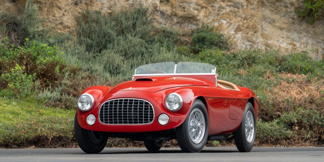 A vintage red convertible sports car with a large front grille and wire-spoke wheels is parked on a paved road with greenery and a rocky background—an icon worthy of Monterey 2025 or the Mecum Auction’s Ferrari Legends.